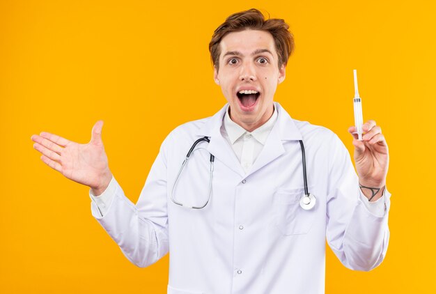 Surprised young male doctor wearing medical robe with stethoscope holding syringe points with hand at side isolated on orange wall