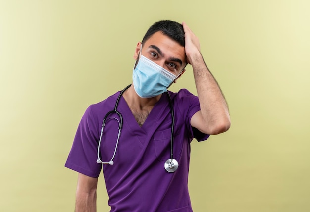 Удивленный молодой врач-мужчина в фиолетовой одежде хирурга и в медицинской маске со стетоскопом положил руку на голову на изолированной зеленой стене