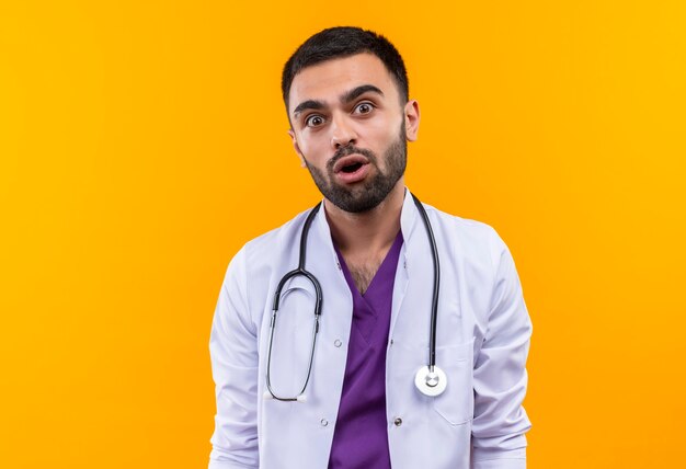 Surprised young male doctor wearing stethoscope medical gown on isolated yellow wall