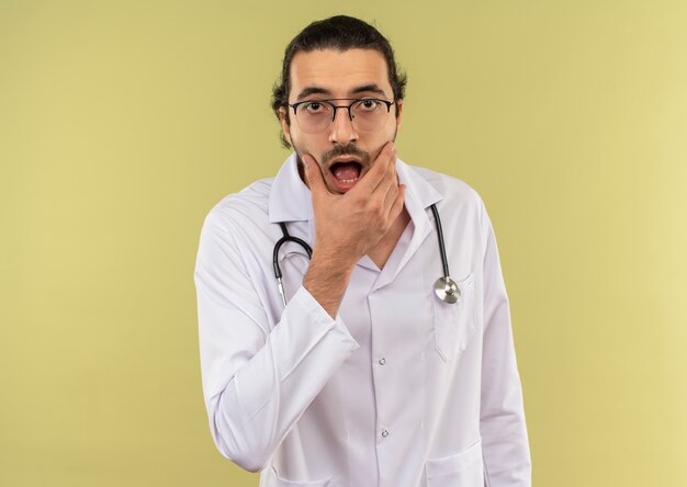 Surprised young male doctor with optical glasses wearing white robe with stethoscope putting hand on chin on isolated green wall with copy space