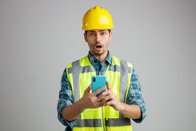 Удивленный молодой инженер-мужчина в защитном шлеме и униформе, держащий мобильный телефон обеими руками и смотрящий в камеру на белом фоне Premium Фотографии