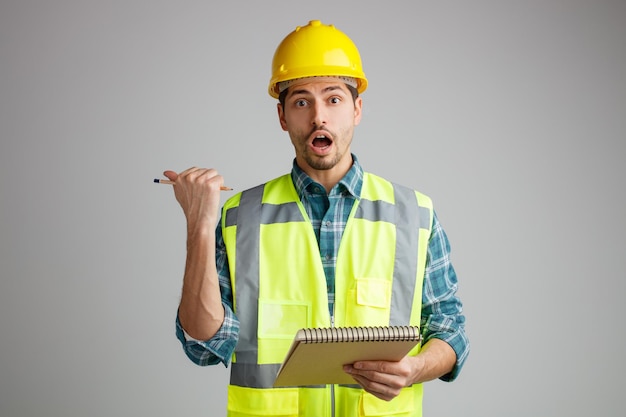 Surprised young male engineer wearing safety helmet and uniform holding note pad and pencil looking at camera pointing to side isolated on white background