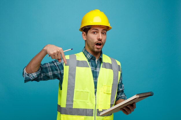 Surprised young male engineer wearing safety helmet and uniform holding pencil and note pad looking at camera pointing down isolated on blue background