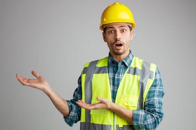 Удивленный молодой инженер-мужчина в защитном шлеме и униформе смотрит в камеру, показывая пустые руки, изолированные на белом фоне
