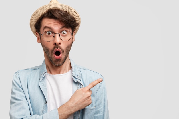 Surprised young male farmer, wears casual shirt and straw hat, has stubble, points at upper right corner