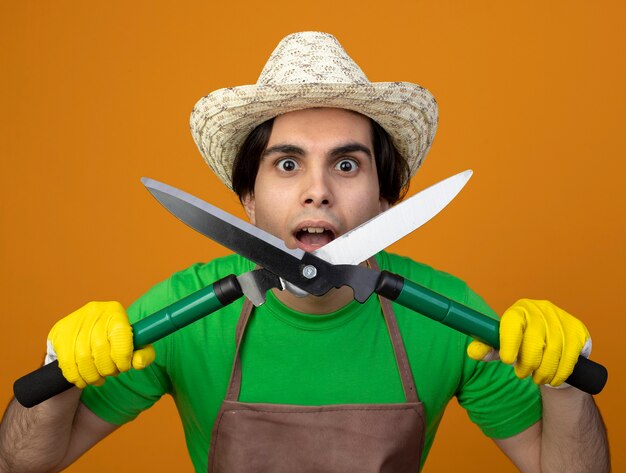 Surprised young male gardener in uniform wearing gardening hat with gloves holding clippers isolated on orange wall