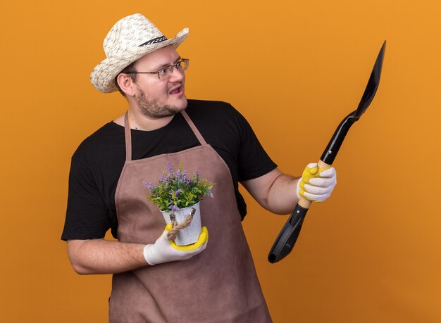 Surprised young male gardener wearing gardening hat and gloves looking at spade in his hand holding flower in flowerpot isolated on orange wall