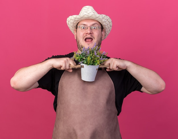 Foto gratuita giovane giardiniere maschio sorpreso che indossa il cappello di giardinaggio che tiene fiore in vaso di fiori isolato sulla parete rosa