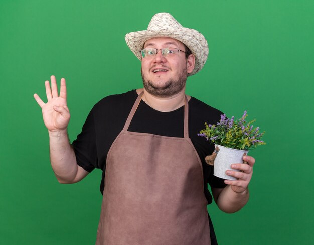 Surprised young male gardener wearing gardening hat holding flower in flowerpot showing four isolated on green wall