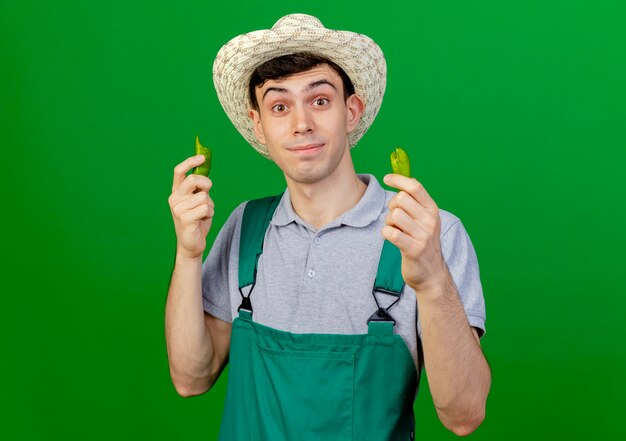 ガーデニング帽子をかぶって驚いた若い男性の庭師は、コピースペースで緑の背景に分離されたカメラを見て壊れた唐辛子を保持します。