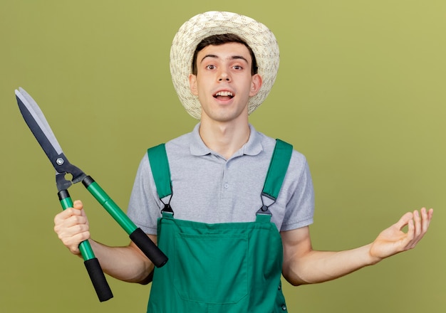 Удивленный молодой мужчина-садовник в садовой шляпе держит машинку для стрижки и держит открытую руку