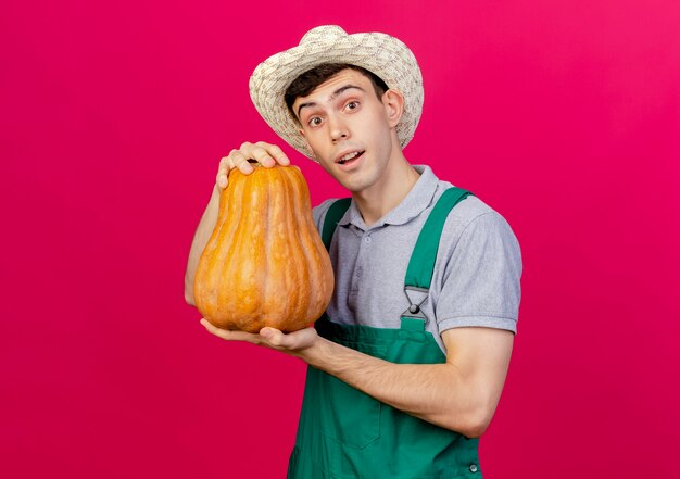 Surprised young male gardener wearing gardening hat holds pumpkin looking at camera