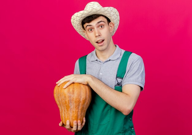 Surprised young male gardener wearing gardening hat holds pumpkin