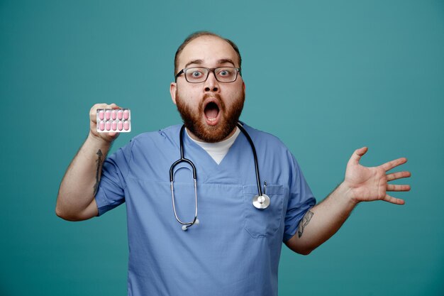 Surprised young male nurse wearing glasses nurse scrub and stethoscope around his neck showing pack of pills and empty hand looking at camera isolated on blue background