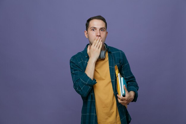 Surprised young male student wearing headphones around neck holding study tools under arm looking at camera making oops gesture isolated on purple background with copy space