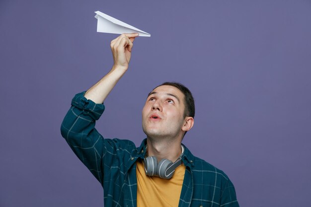 紫色の背景に分離されたそれを見て頭上に紙飛行機を上げる首の周りにヘッドフォンを身に着けている驚いた若い男子学生