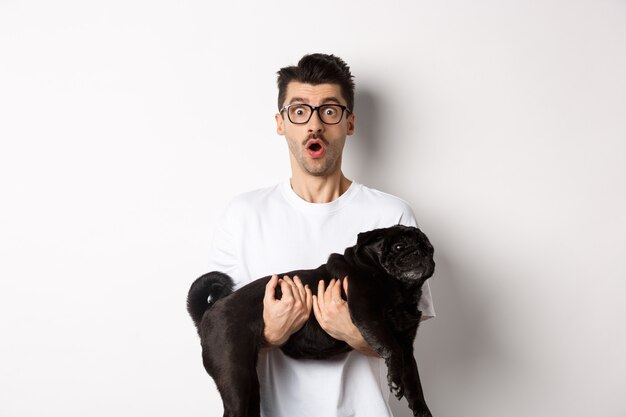 Surprised young man in glasses holding cute black pug, dog owner staring at camera with impressed face, saying wow, standing over white background