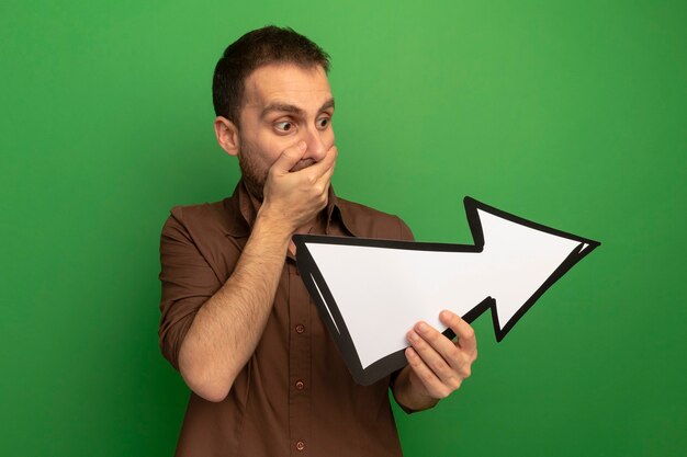 Surprised young man holding and looking at arrow mark pointing at side keeping hand on mouth isolated on green wall