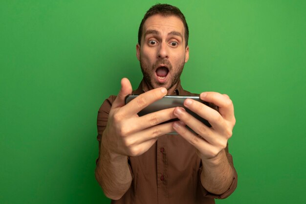 Surprised young man holding and looking at mobile phone isolated on green wall