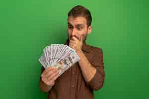 Free photo surprised young man holding and looking at money keeping hand on mouth isolated on green wall