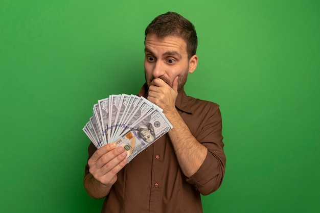 Удивленный молодой человек, держащий и смотрящий на деньги, держа руку на рту, изолированный на зеленой стене