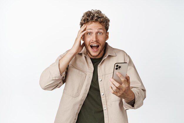 Surprised young man holding mobile phone looking in disbelief at camera amazed by smth on smartphone app standing against white background