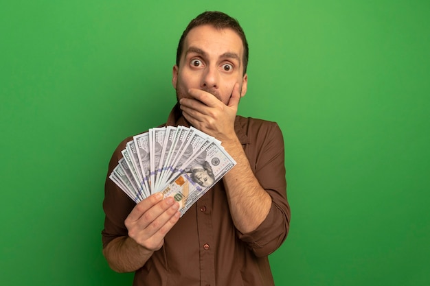 Surprised young man holding money looking at front putting hand on mouth isolated on green wall