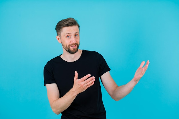 Surprised young man is pointing right by raising up hand to rigth on blue background