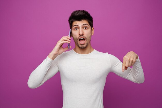 Free photo surprised young man looking at camera pointing down while talking on phone isolated on purple background