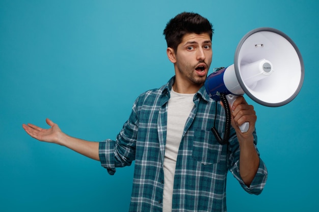 Free photo surprised young man looking at camera talking into speaker showing empty hand isolated on blue background