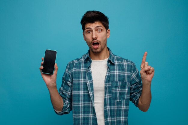 Удивленный молодой человек показывает мобильный телефон камере, глядя на камеру, указывающую вверх на синем фоне