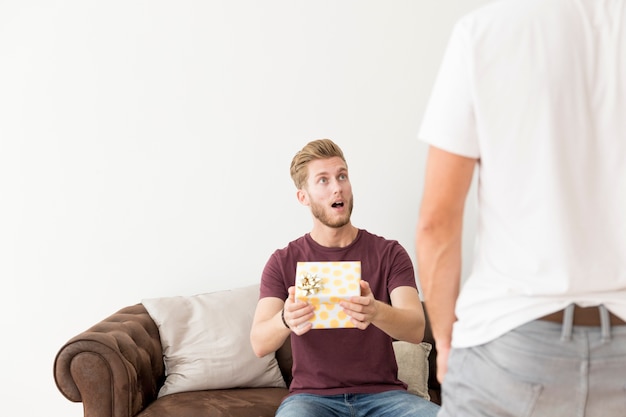 Foto gratuita sorpreso giovane uomo seduto sul divano tenendo scatola regalo guardando il suo amico