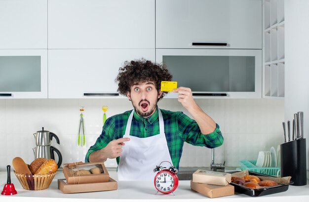 Surprised young man standing behind the table various pastries on it and showing bank card in the white kitchen