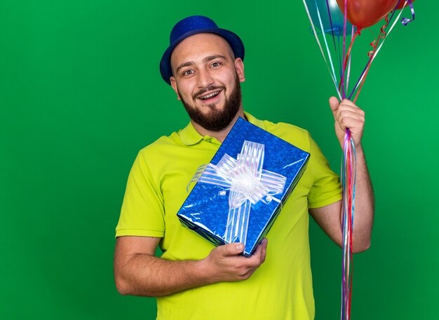 Удивленный молодой человек в синей партийной шляпе держит воздушные шары с подарочной коробкой