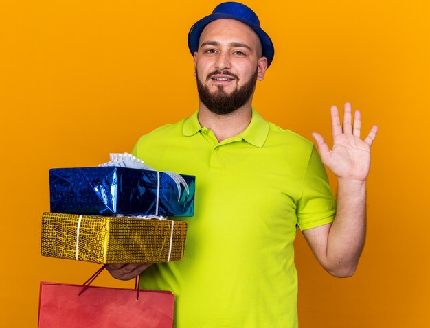 Surprised young man wearing party hat holding gift boxes with bag showing hello gesture isolated on orange wall