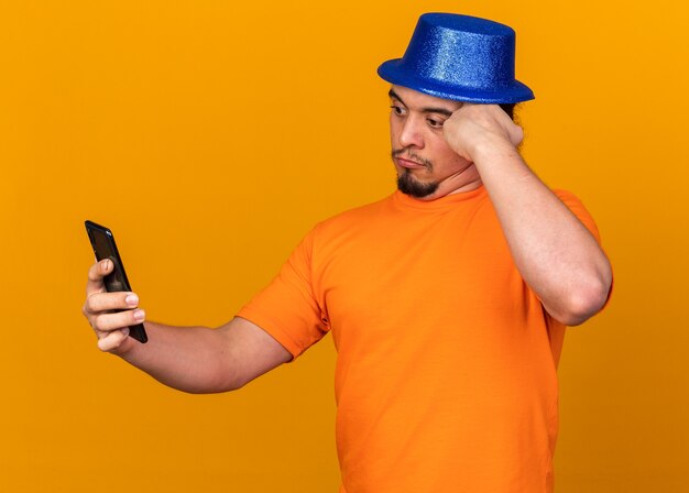 Удивленный молодой человек в партийной шляпе, держащий и смотрящий в телефон, положив руку на храм, изолированный на оранжевой стене