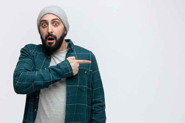 Surprised young man wearing winter hat looking at camera pointing to side isolated on white background with copy space