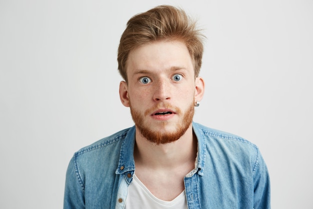 Surprised young man with beard in jean shirt looking stretching to camera.