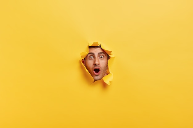 Free photo surprised young man with bristle keeps mouth widely opened, stares through torn paper wall