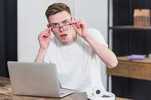 Surprised young man with headphone and laptop on wooden table looking to camera