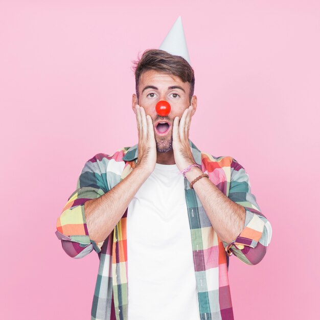 Surprised young man with red clown nose standing against pink backdrop