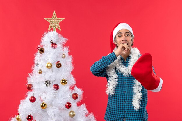 青い縞模様のシャツを着て、彼のクリスマスの靴下を身に着けているサンタクロースの帽子をかぶった驚いた若い男