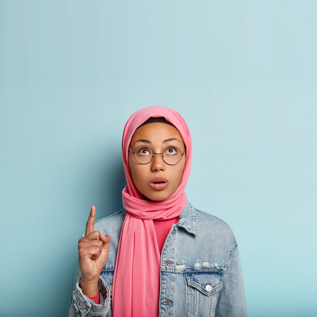 Surprised young Muslim woman points up with one fore finger, focused upwards, has concerned facial expression, veiled head