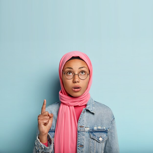 Surprised young Muslim woman points up with one fore finger, focused upwards, has concerned facial expression, veiled head