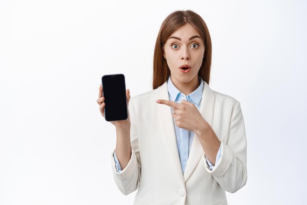 Surprised young office girl in business suit gasps and points left at smartphone screen shows online promo offer white background