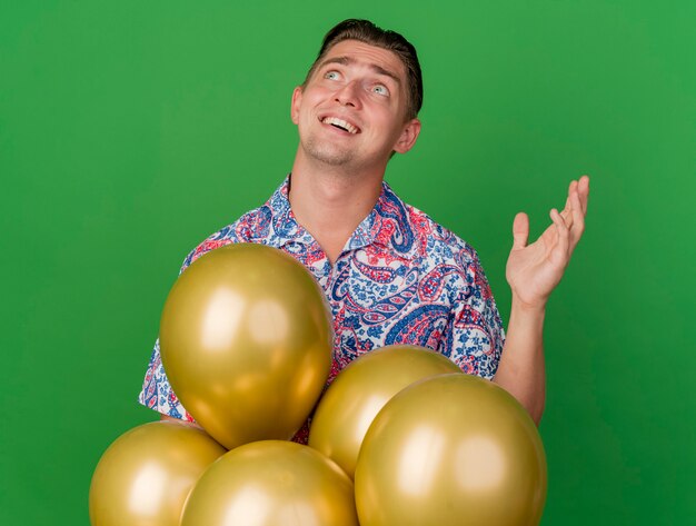 Surprised young party guy looking at up wearing colorful shirt standing behind balloons raising hand isolated on green