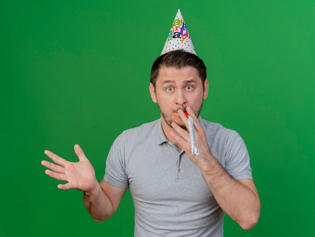 surprised young party guy wearing birthday cap holding and blowing whistle spreading hand isolated on green