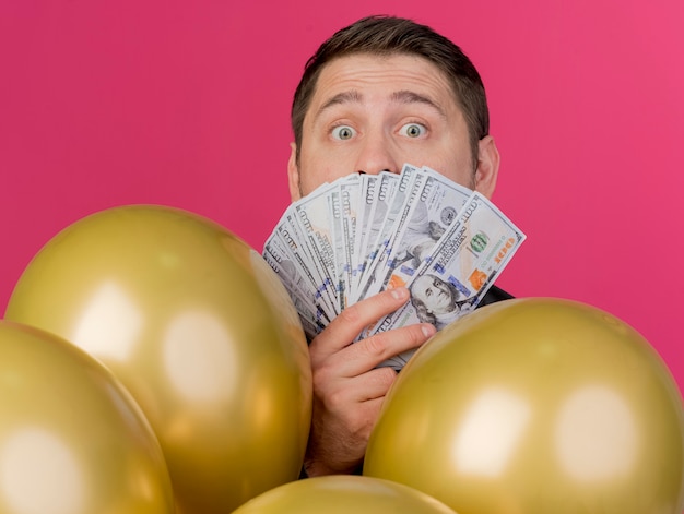 Surprised young party guy wearing black shirt standing behind balloons covered face with cash isolated on pink