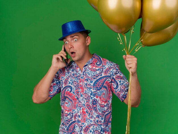 Удивленный молодой тусовщик в синей шляпе с воздушными шарами разговаривает по телефону на зеленом фоне