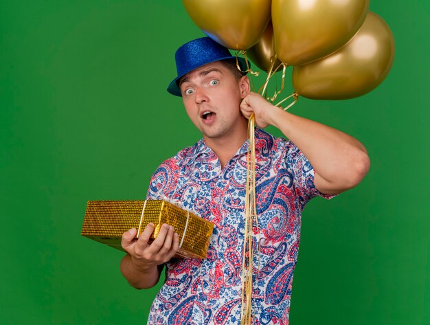 녹색에 고립 된 풍선과 함께 선물 상자를 들고 파란색 모자를 쓰고 놀란 된 젊은 파티 남자